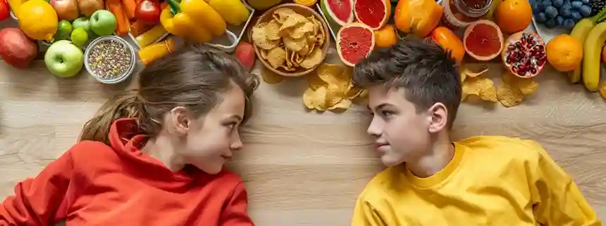 Teen boy and girl looking at a split table with healthy foods like fruits, vegetables, and whole grains on one side, and unhealthy options like chips, soda, and candy on the other.