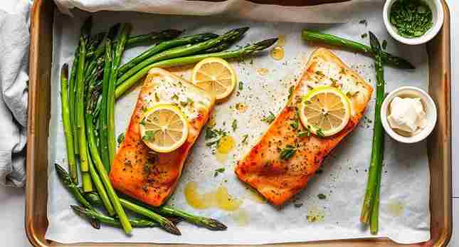 Baked salmon with asparagus on a parchment-lined pan, garnished with lemon slices and herbs.