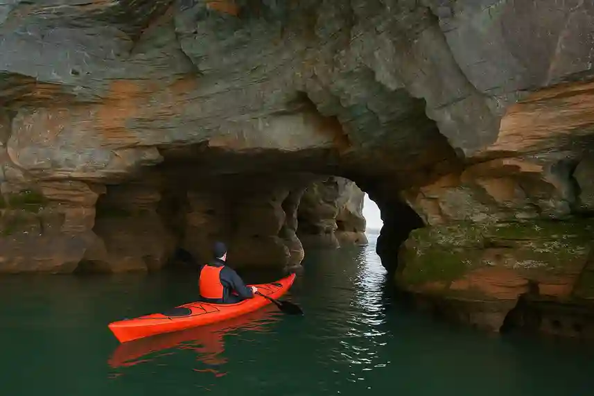 A kayaker paddling through the sea caves of the Apostle Islands, surrounded by red sandstone cliffs and calm green waters.